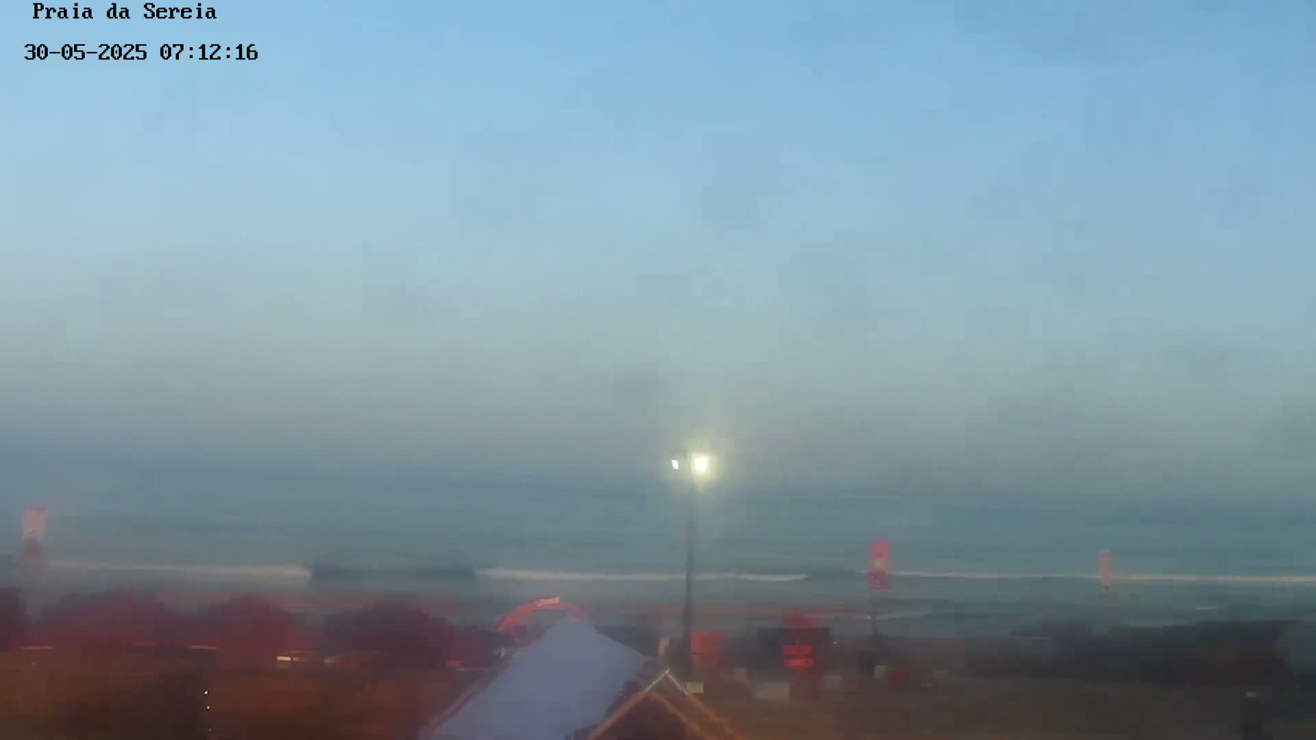 A hazy sunrise illuminates a calm ocean and beach scene with several structures visible near the shoreline.