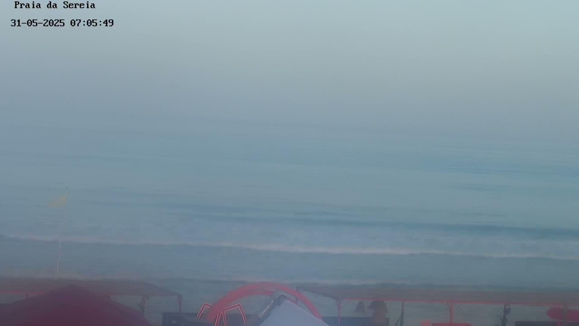 A hazy, overcast day at a beach shows calm waves lapping the shore, with beach umbrellas and some beach-goers visible in the foreground.