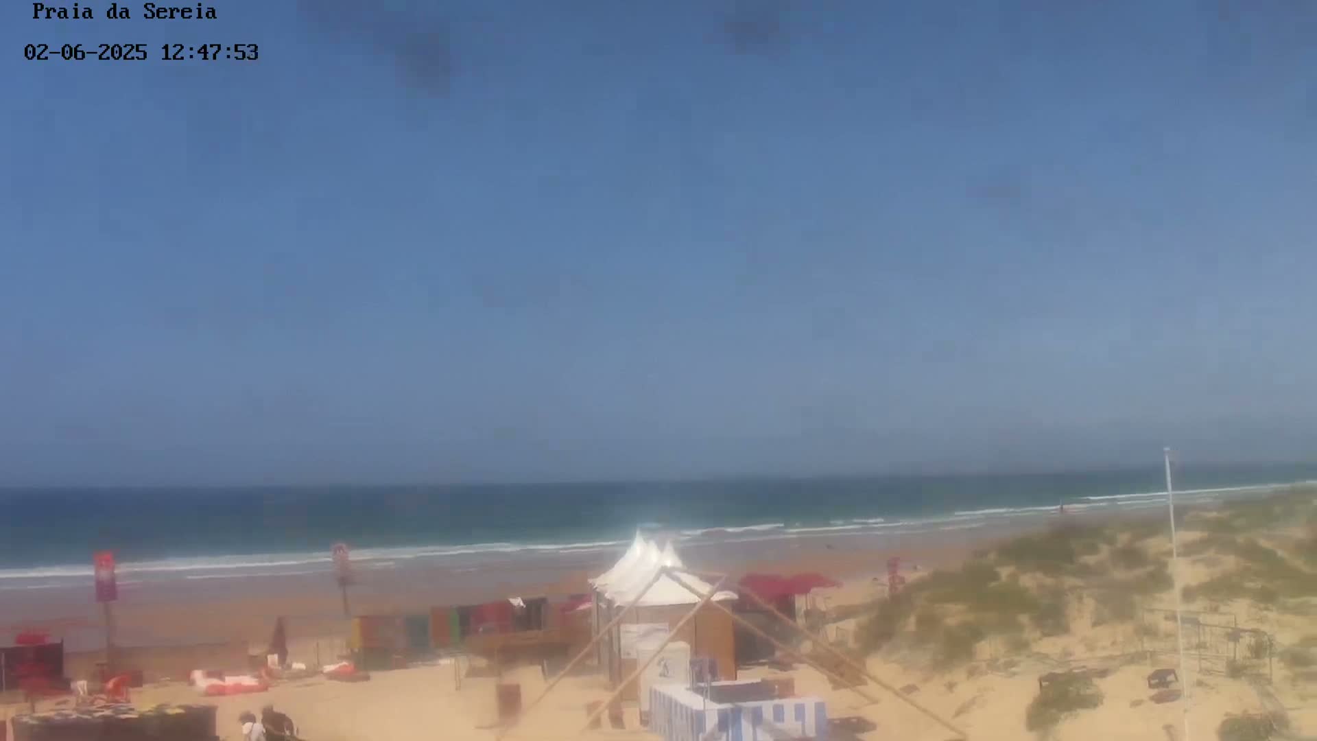 A sandy beach with small structures and a few people is visible under a clear blue sky, with calm ocean waves rolling onto the shore.