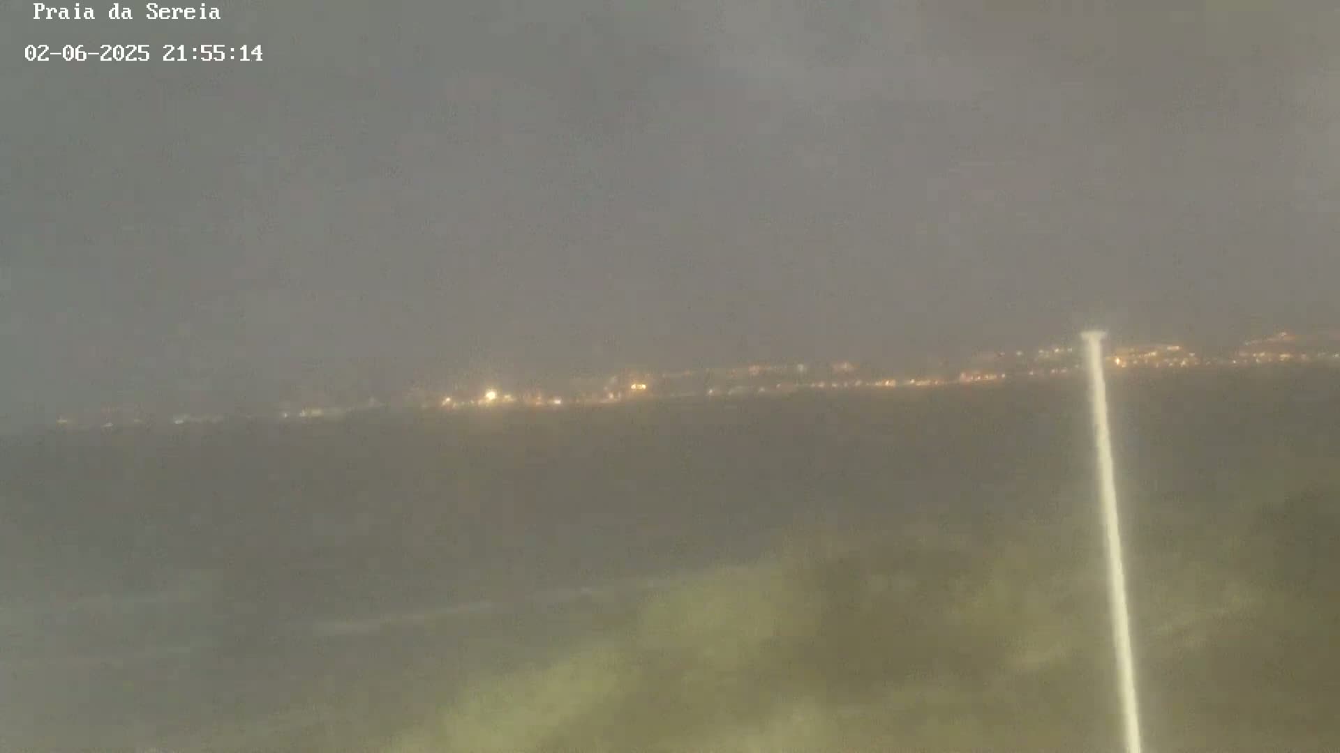 A hazy night scene shows a distant line of lights, possibly a town, with a bright vertical light in the foreground, possibly a light source.
