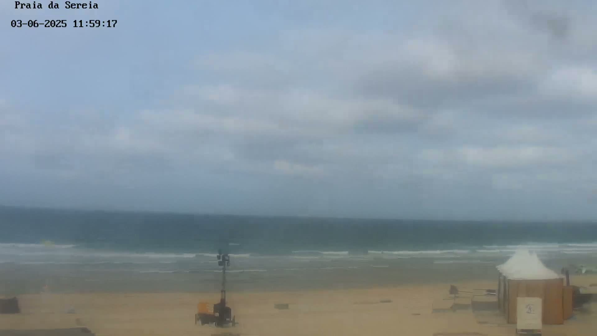 A mostly cloudy day shows an ocean beach with small waves, a tall piece of equipment, and a small beach hut.