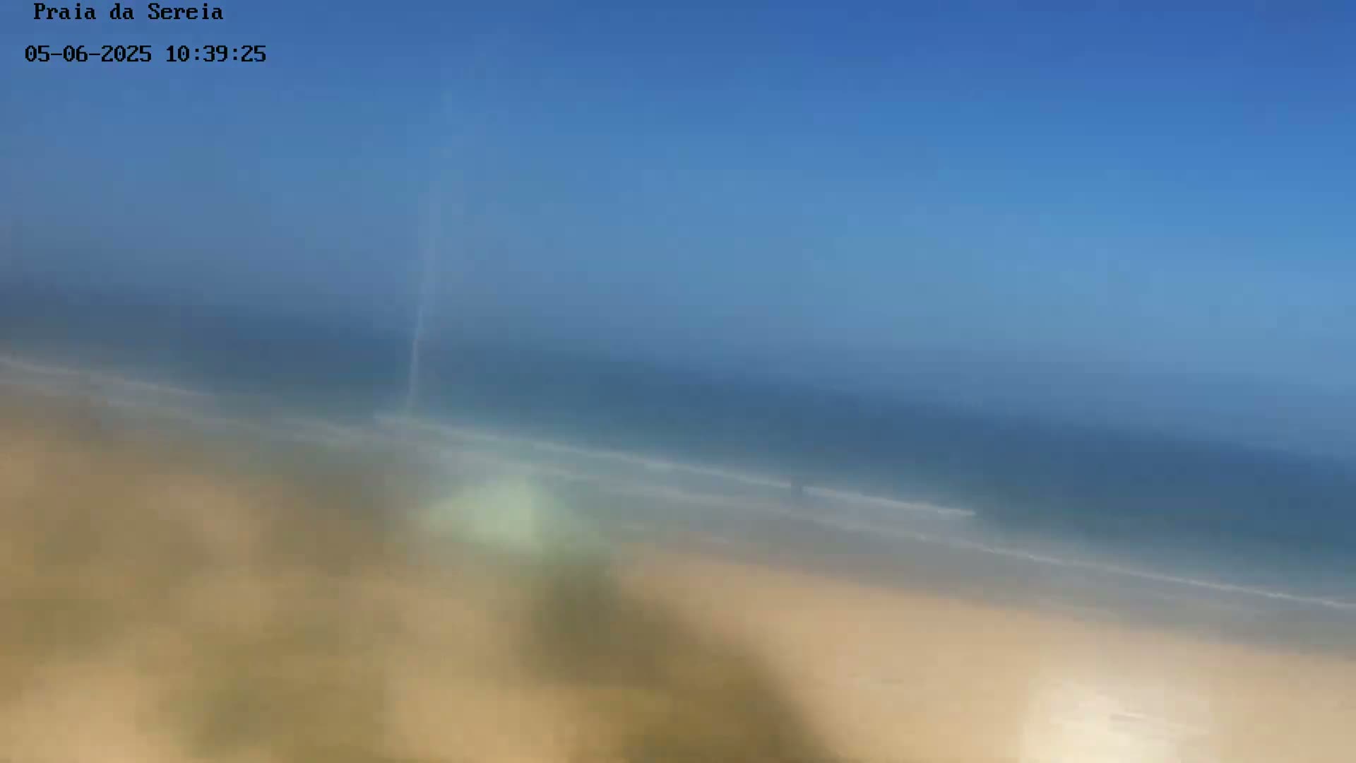 A hazy, sunny day shows a sandy beach meeting a calm ocean under a clear blue sky.