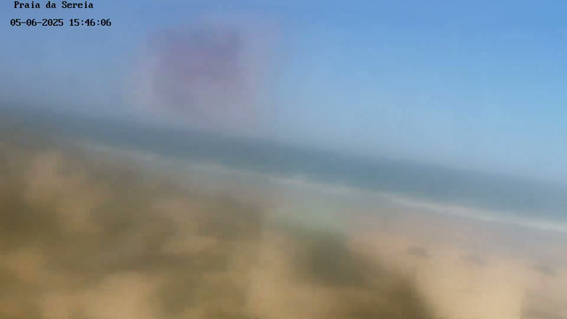 A blurry image shows a sandy beach meeting a calm ocean under a clear blue sky.