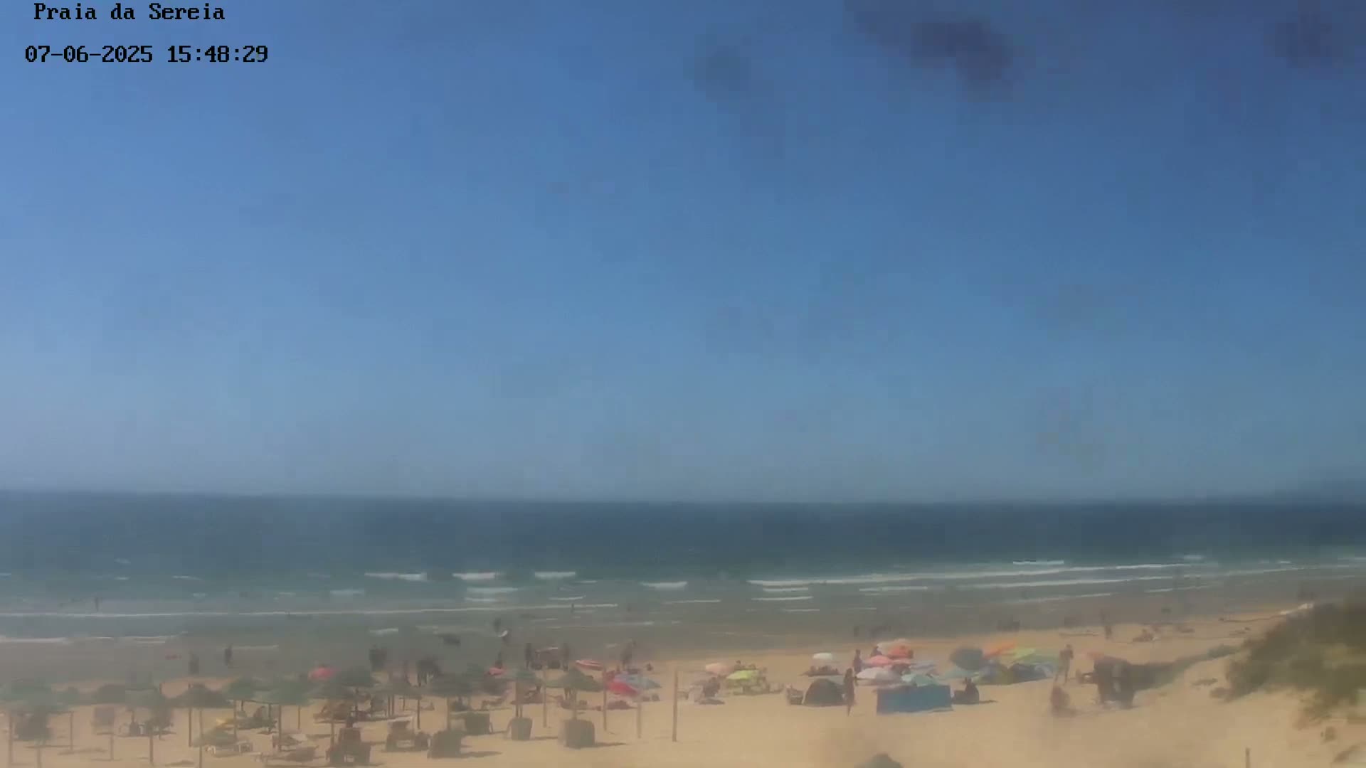A sandy beach with people and umbrellas under a clear blue sky and a calm ocean with small waves.