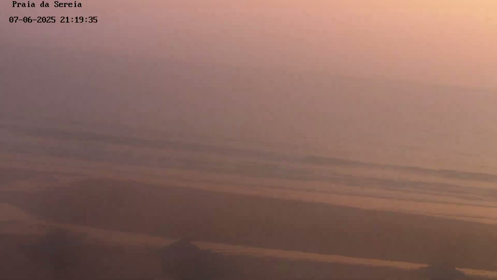 A hazy, pinkish-orange sunset illuminates a beach and ocean with gentle waves.
