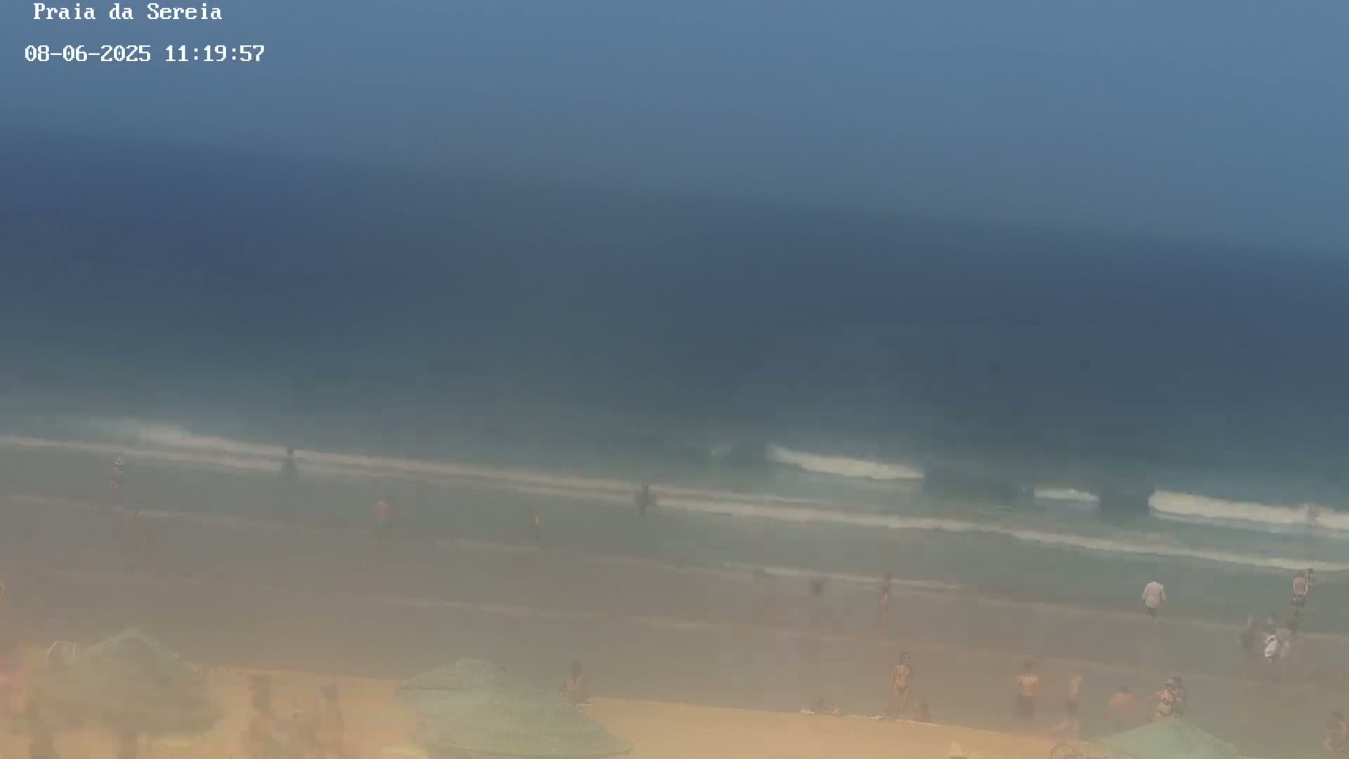 A hazy beach scene shows people swimming and relaxing under umbrellas on a sandy shore with gentle waves rolling in under a mostly clear sky.