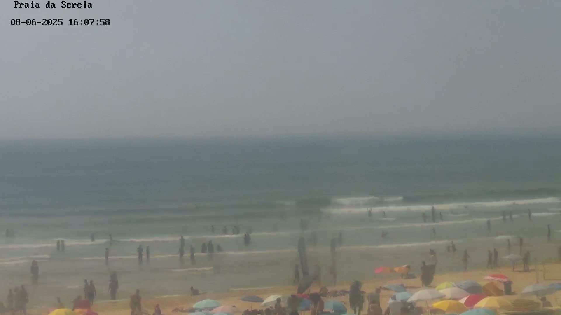 A hazy beach scene shows many people swimming and sunbathing on a sandy shore with small waves and numerous colorful umbrellas.