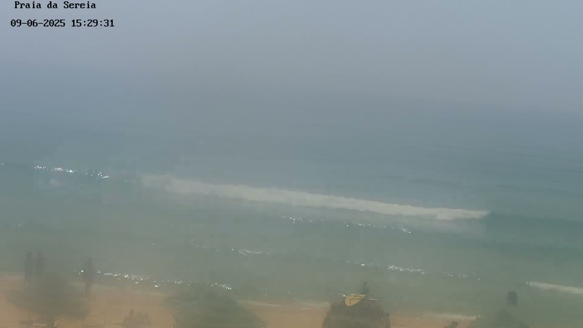 A hazy, overcast day at a beach shows small waves lapping the shore, with a few people visible on the sand.
