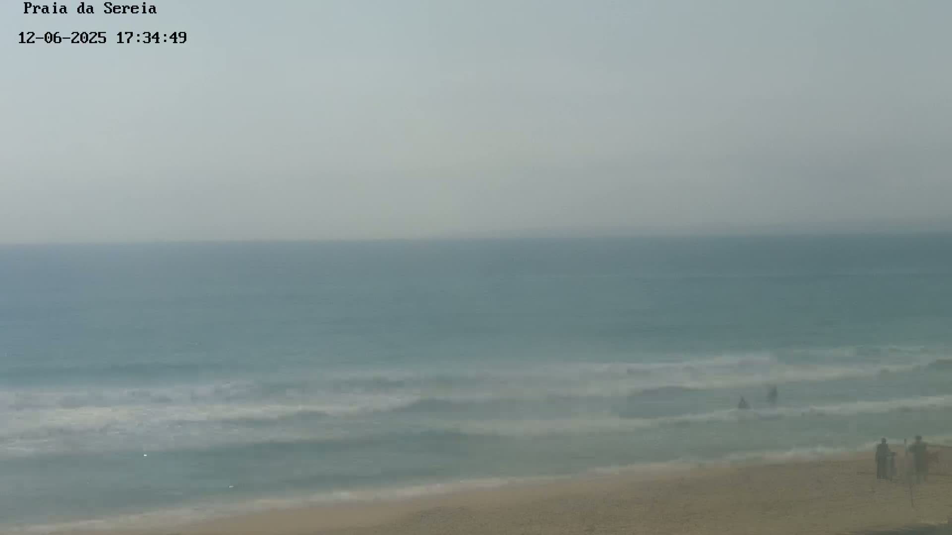A hazy view shows a calm ocean with small waves lapping a sandy beach where a few people stand.