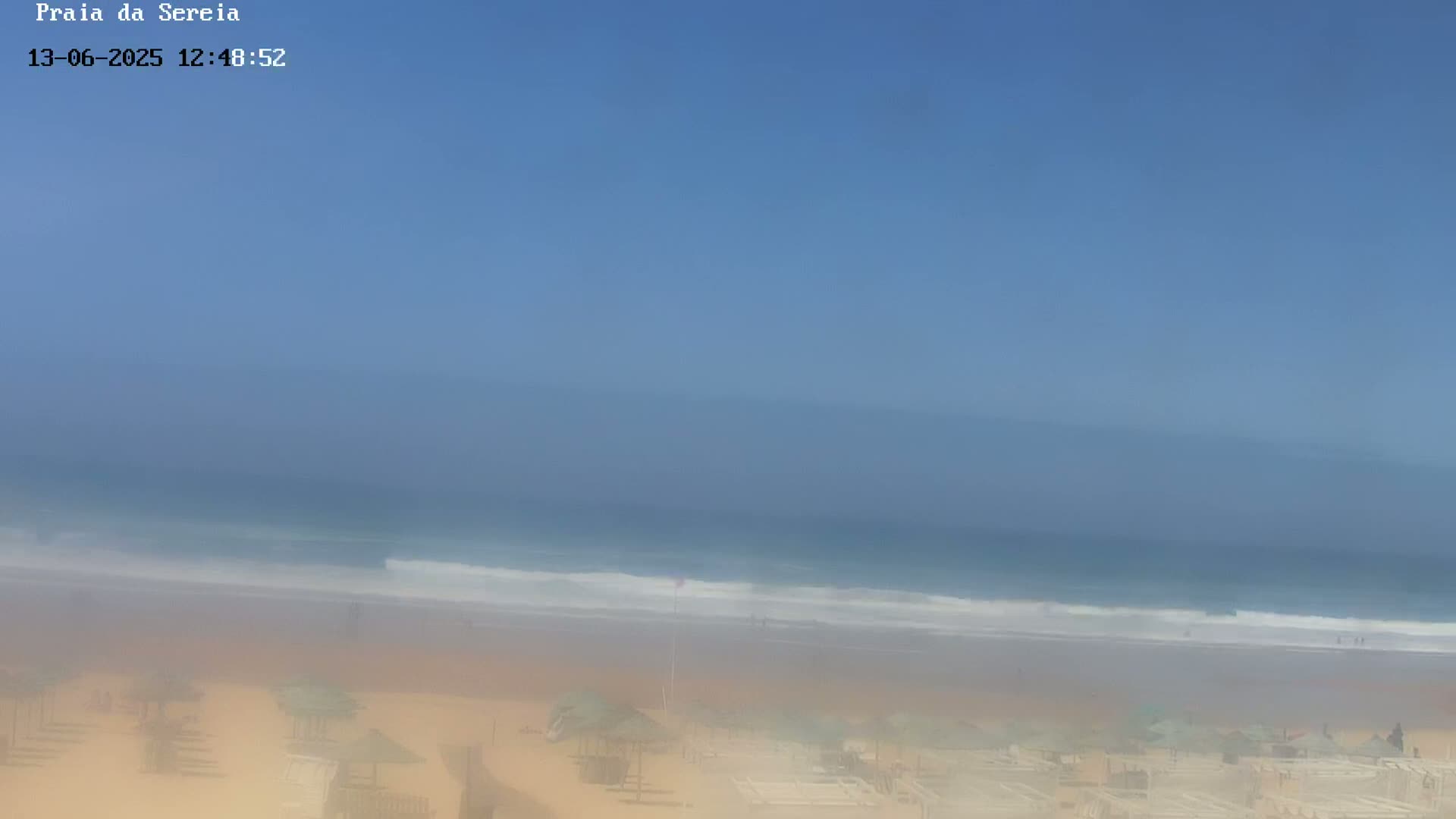 A hazy, sunny day at a beach shows a calm ocean with small waves breaking on the shore, where there are several beach umbrellas and people visible in the distance.