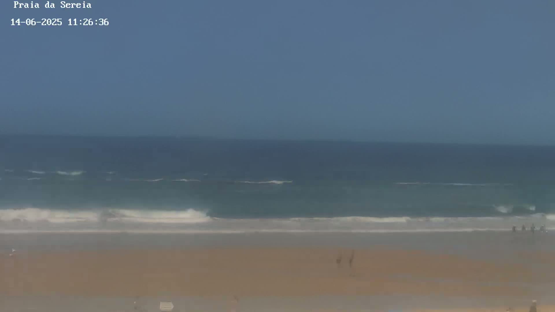 A mostly clear sky overlooks a sandy beach with small waves lapping the shore, and a few people are visible in the distance.