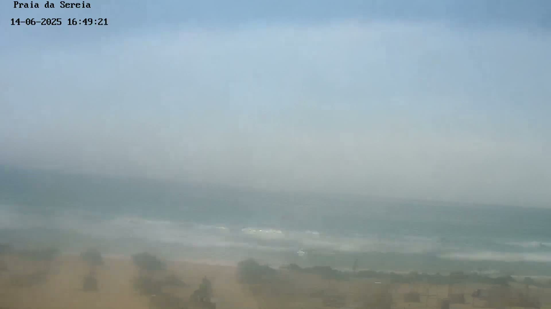 A hazy, misty view shows a beach with waves crashing on the shore under a mostly clear sky.