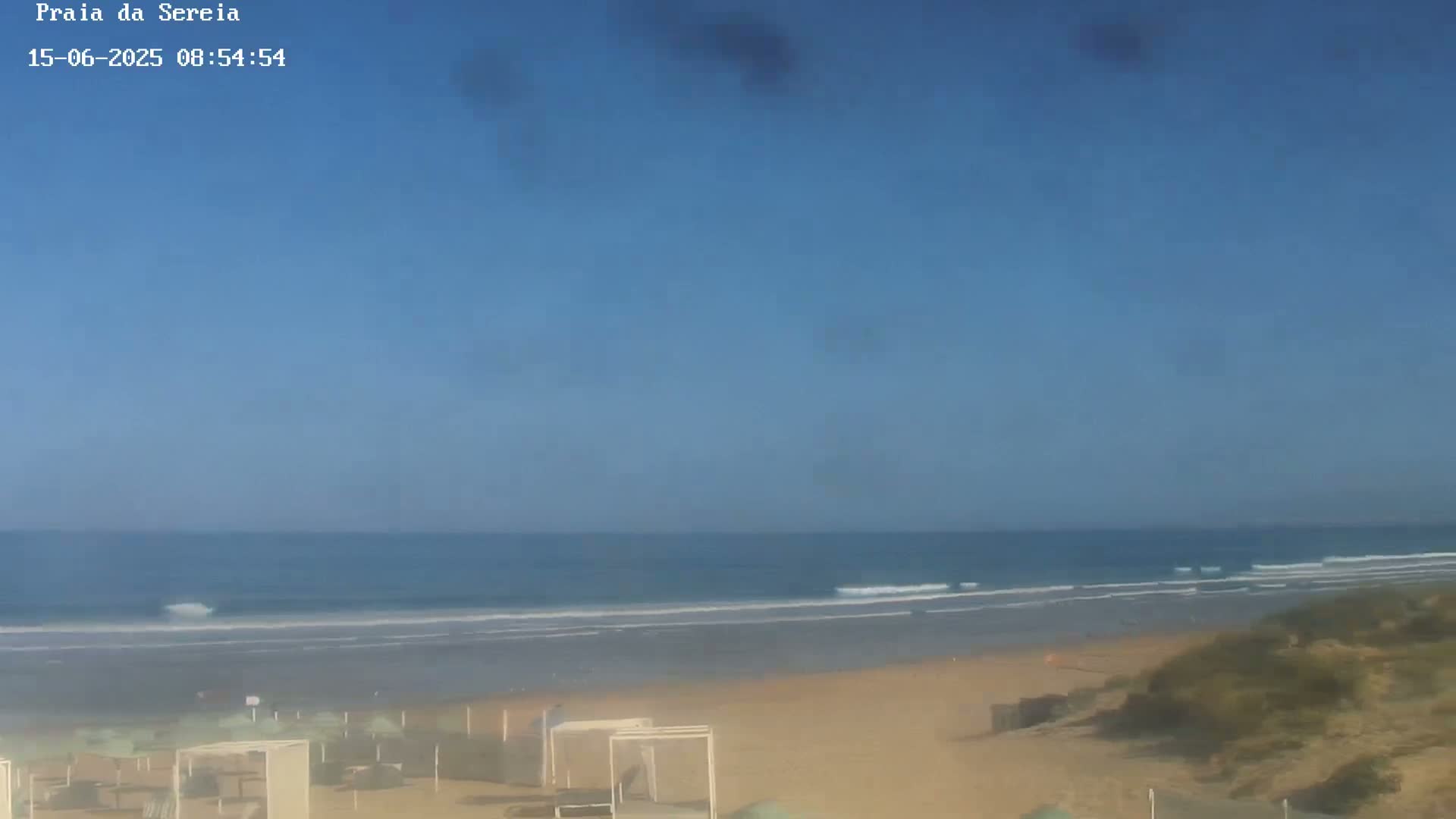 A sandy beach with calm ocean waves under a clear blue sky is shown with several beach umbrellas and some dunes.