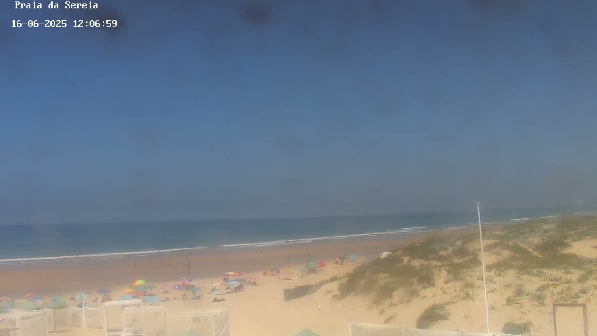 A sandy beach with scattered beach umbrellas and people, overlooking a calm ocean under a clear blue sky.