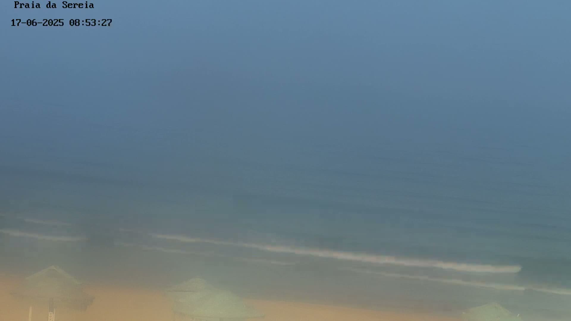 A hazy beach scene shows a calm ocean with small waves gently lapping the shore near several beach umbrellas.