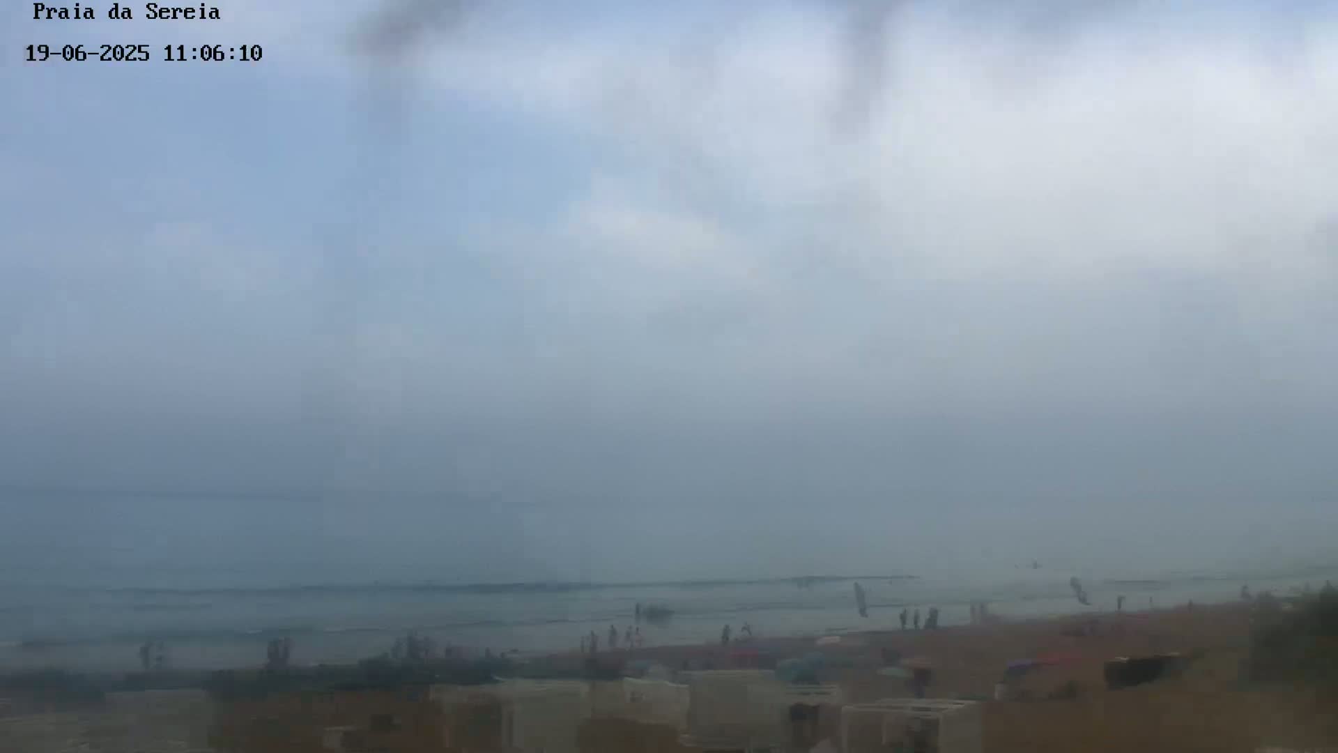 A hazy, overcast day at a beach shows a calm ocean with a few people visible on the sand and in the water.