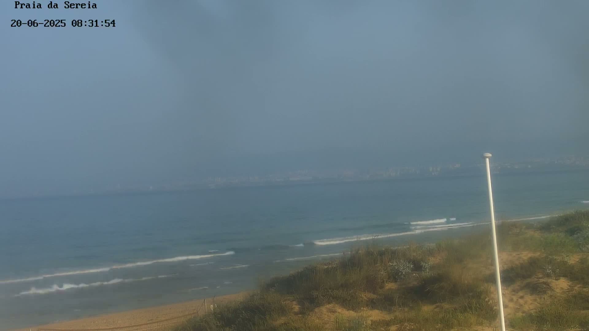 A hazy view of a sandy beach and ocean with small waves, a distant cityscape visible on the horizon.