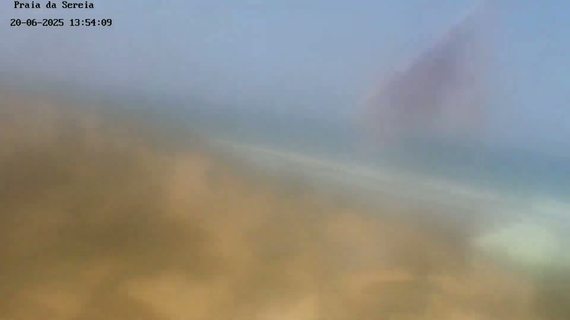 A hazy, blurry view of a sandy beach meeting a calm, light-blue ocean under a pale sky.