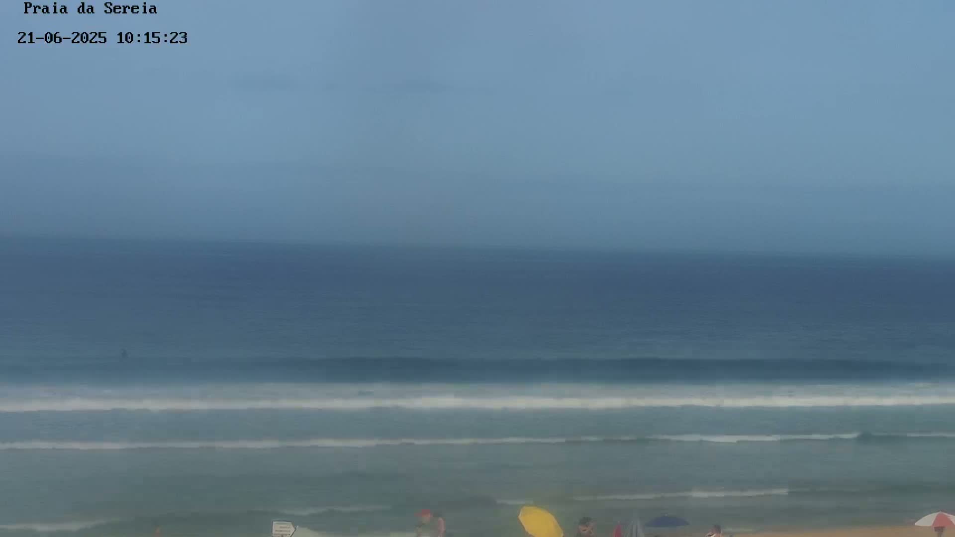 A hazy, overcast day at a beach shows gentle waves rolling onto the shore where a few people are standing near umbrellas.