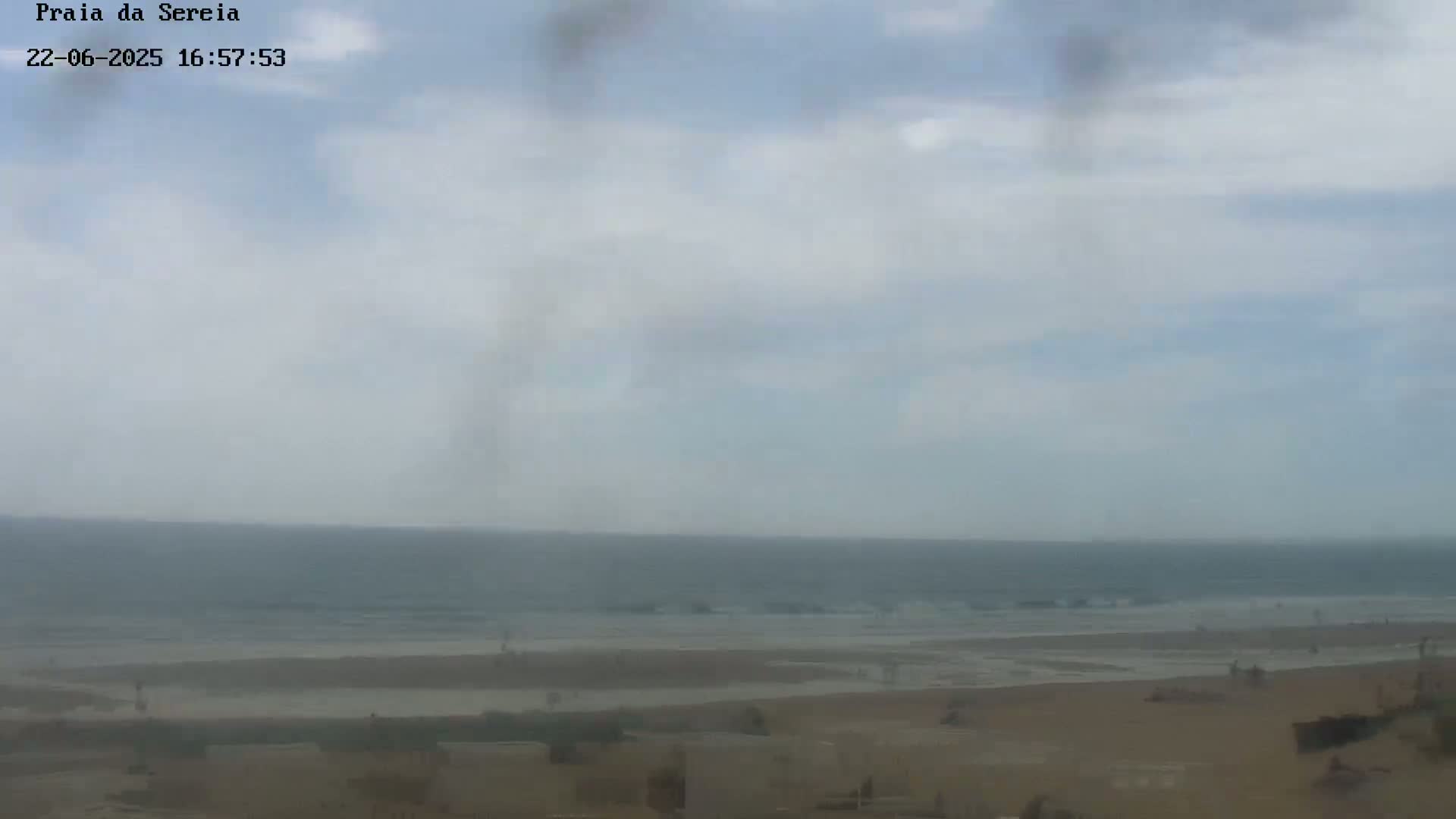 A hazy beach scene shows a calm ocean meeting a sandy shore with a few people visible on the beach under a mostly cloudy sky.