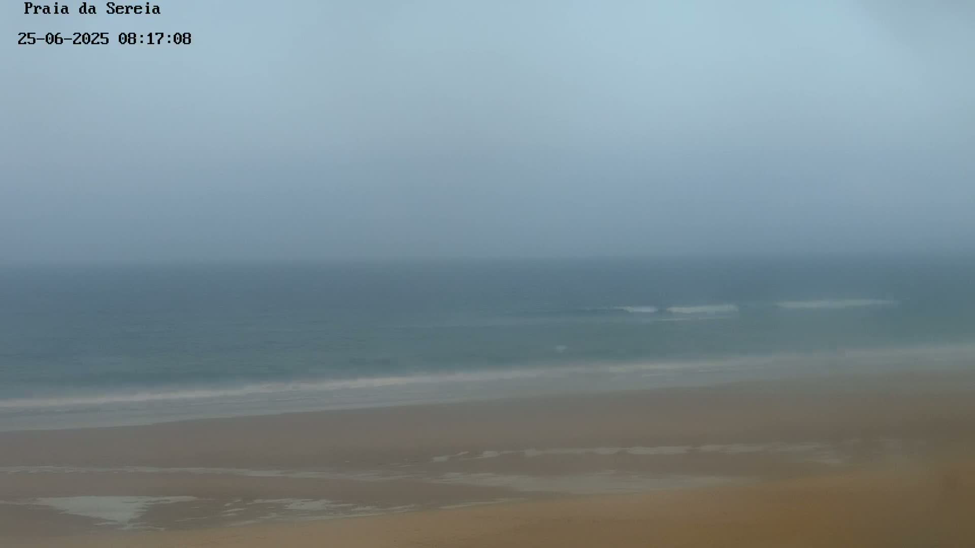 A hazy, overcast day shows a wide expanse of beach and ocean, with small waves visible near the shore.