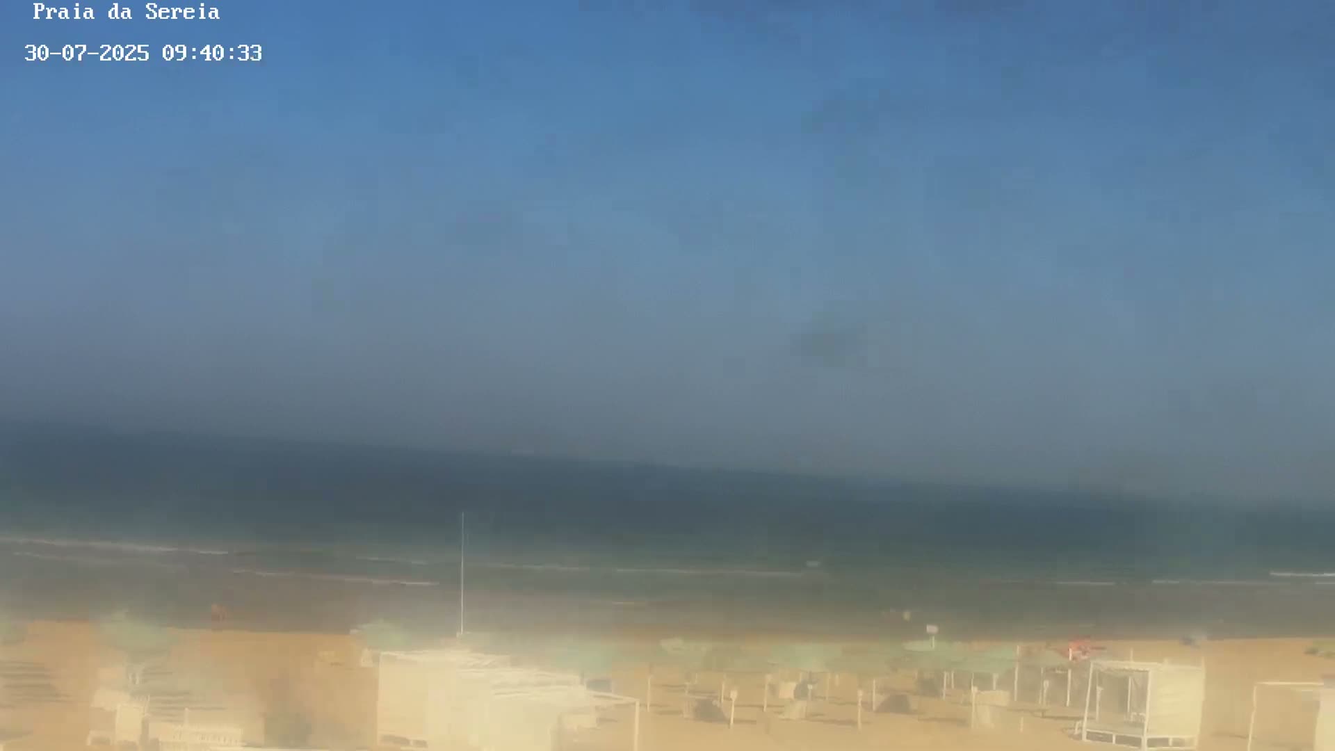 A hazy, sunny day at a beach shows calm ocean waves lapping a sandy shore lined with beach umbrellas and small structures.