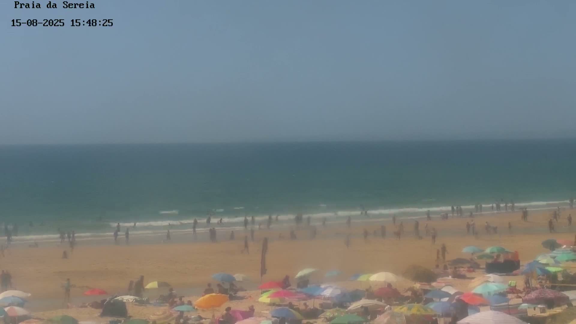 A hazy, sunny day at a beach shows calm ocean waves lapping a sandy shore lined with beach umbrellas and small structures.