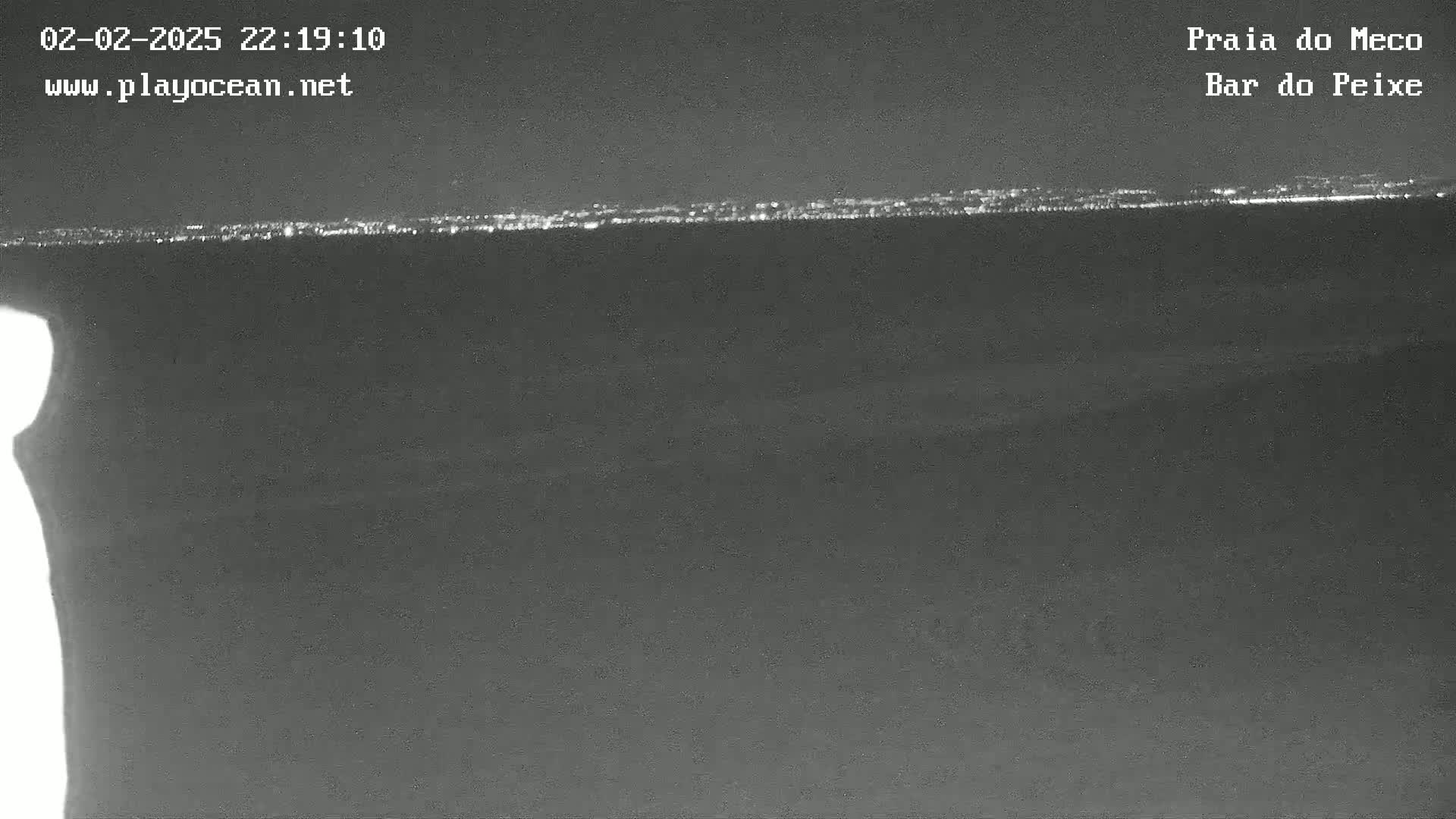 A nighttime, low-angle view shows a dark body of water reflecting a distant line of city lights under a dark sky.
