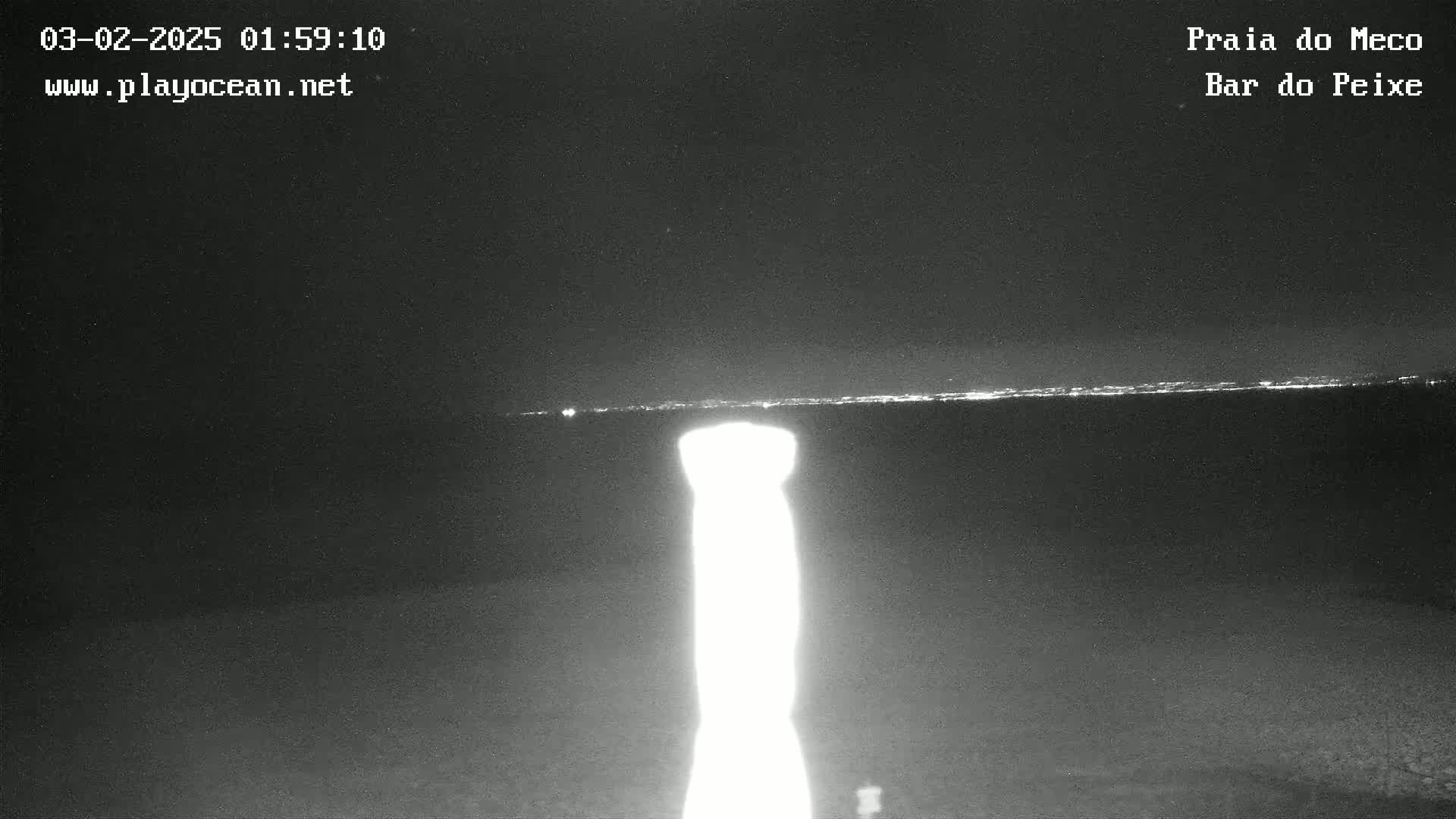 A nighttime beach scene shows a bright light in the foreground and a faint, distant horizon line of city lights.
