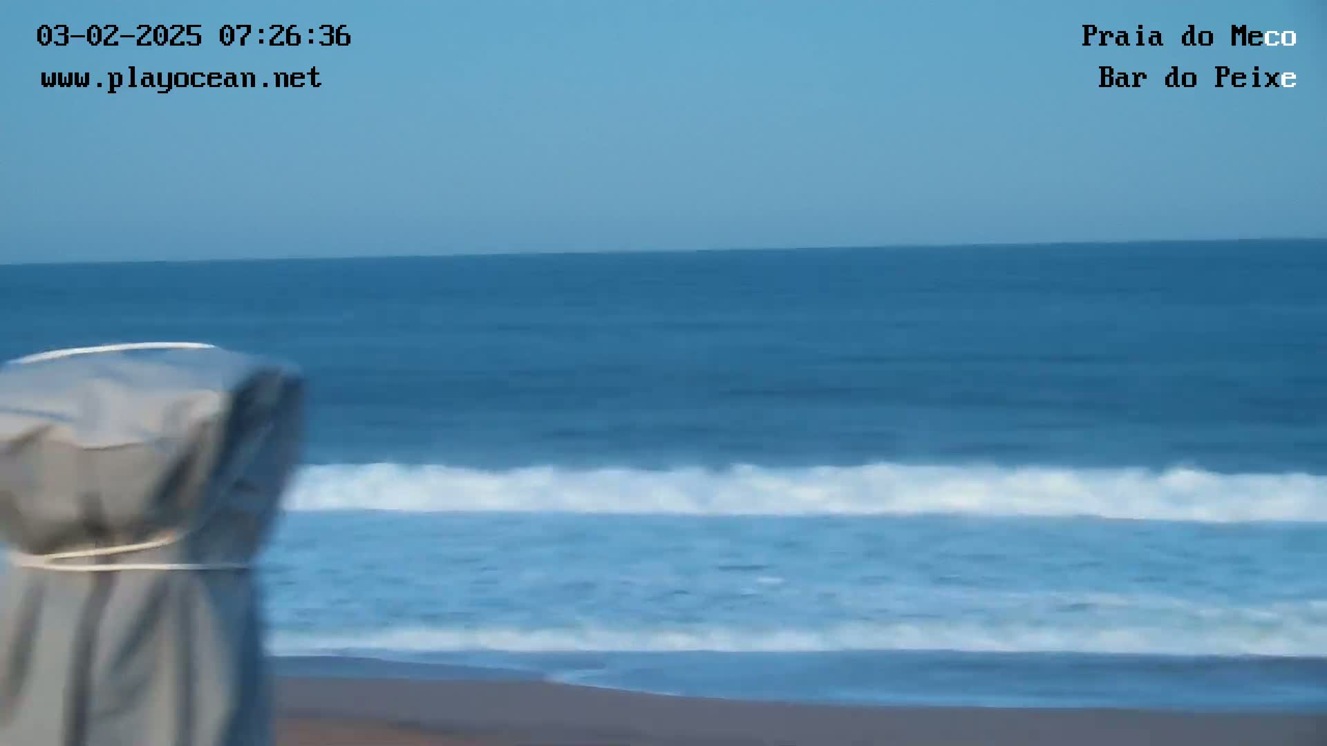 A covered beach umbrella stands on a sandy beach next to a calm ocean under a clear sky.