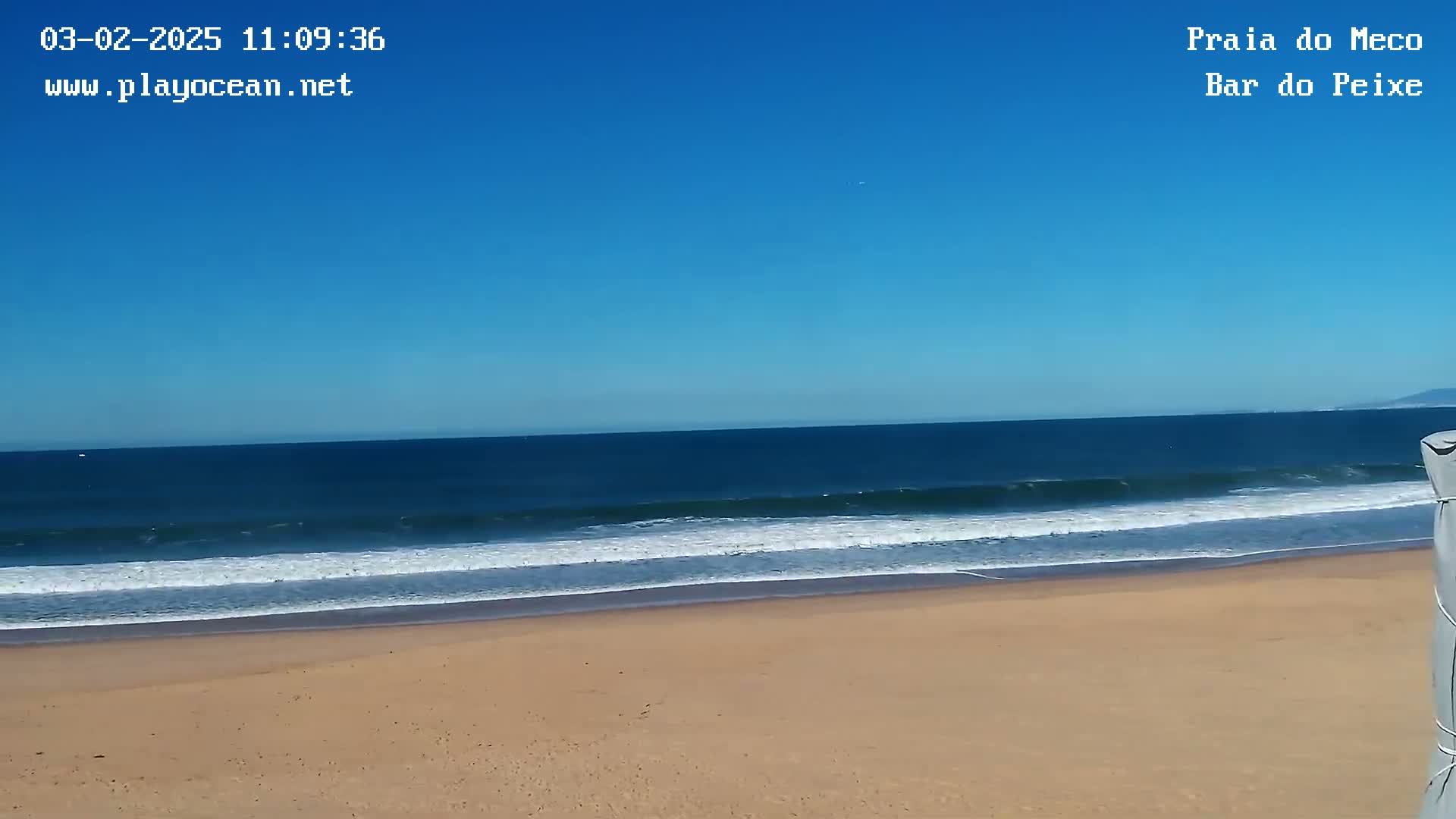 A sandy beach with gentle waves rolls into a calm, blue ocean under a clear sky.