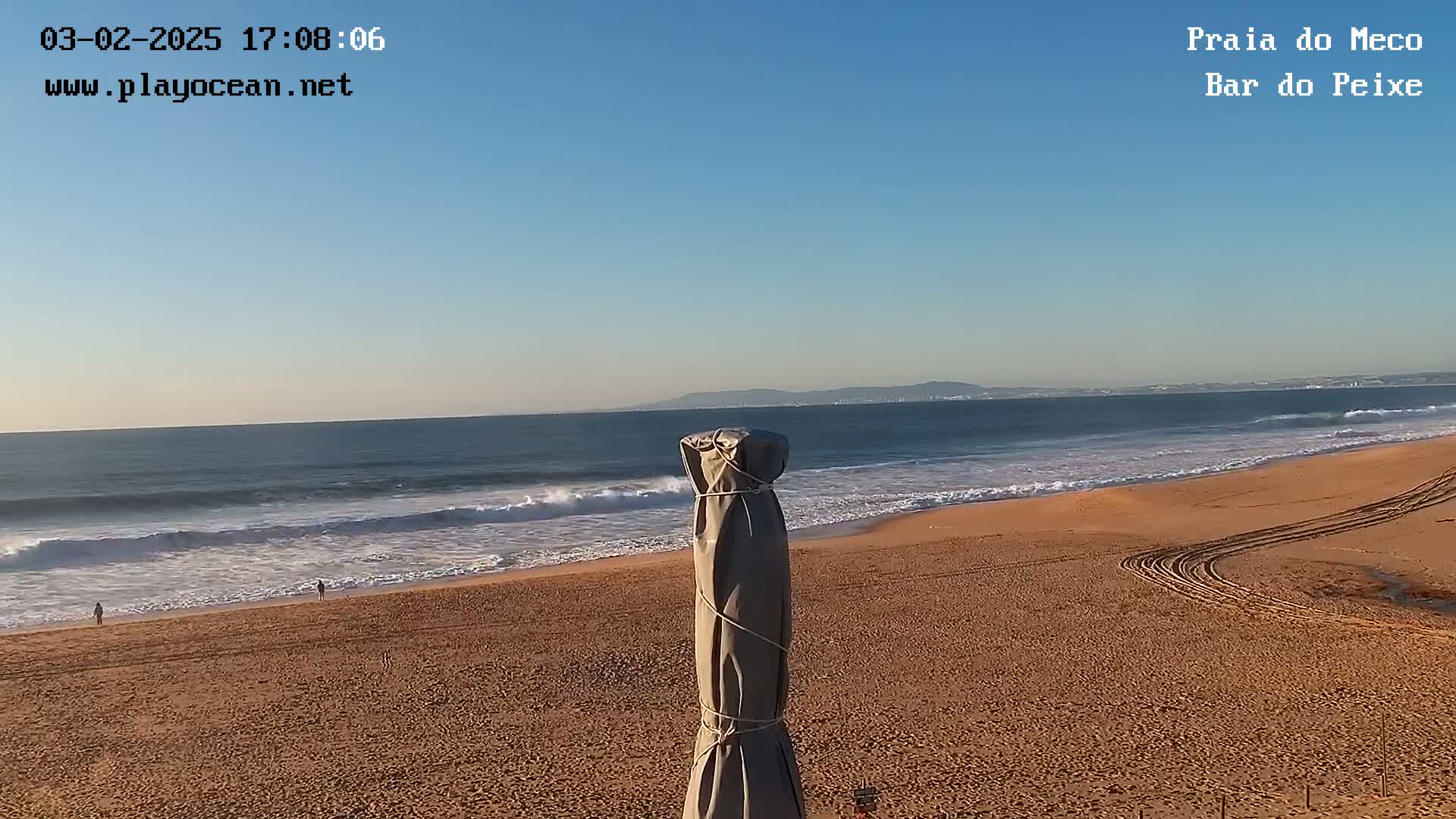 A sandy beach with gentle waves under a clear blue sky, a covered beach umbrella in the foreground, and distant hills visible on the horizon.