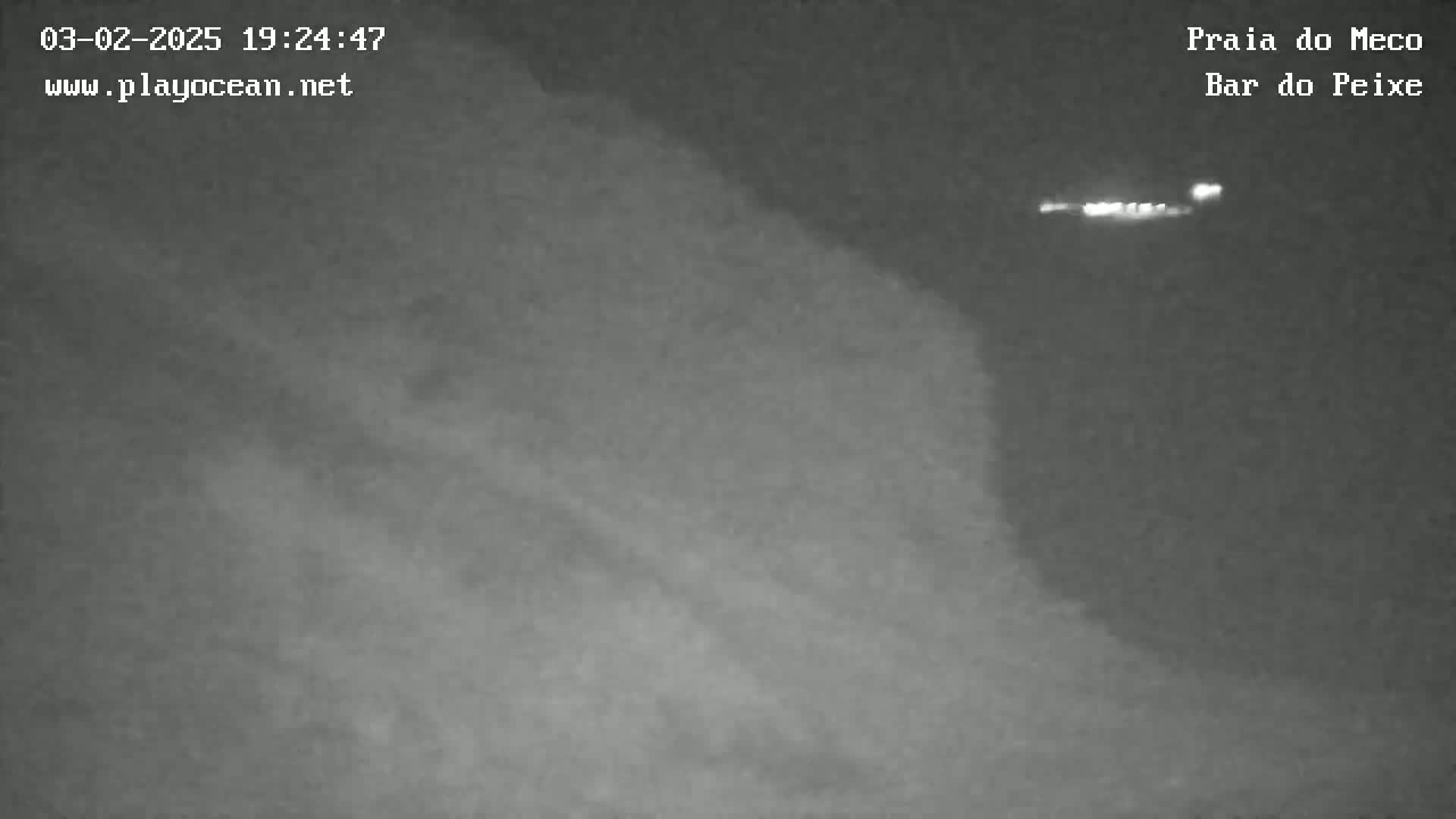 A nighttime, low-light image shows a dark, blurry coastal scene with a faint light source in the distance.