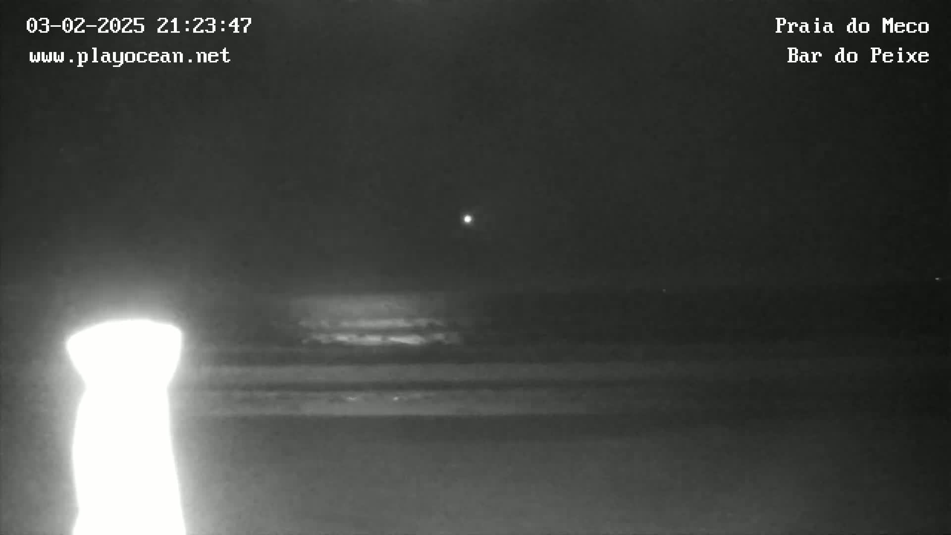 A nighttime, low-light view of a beach with a bright light source in the foreground and a dark, mostly overcast sky.