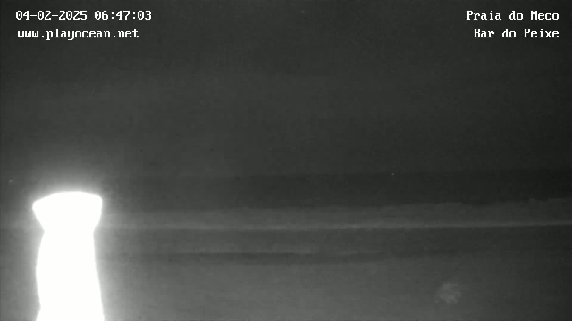 A bright light illuminates the left side of a nighttime, dark beach scene with a calm ocean in the background.