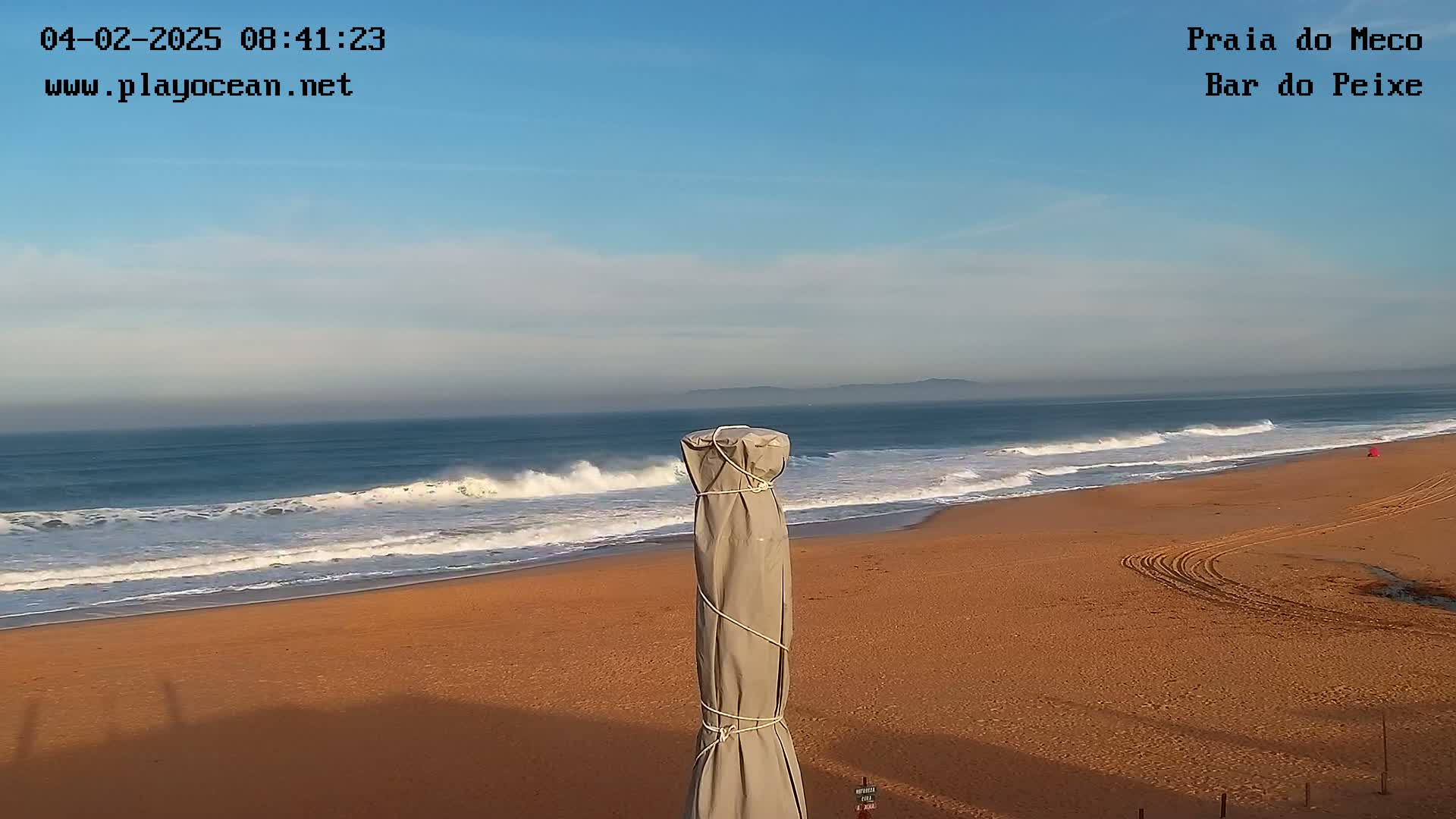 A sandy beach with waves rolling onto the shore under a mostly clear sky, and a covered beach umbrella in the foreground.
