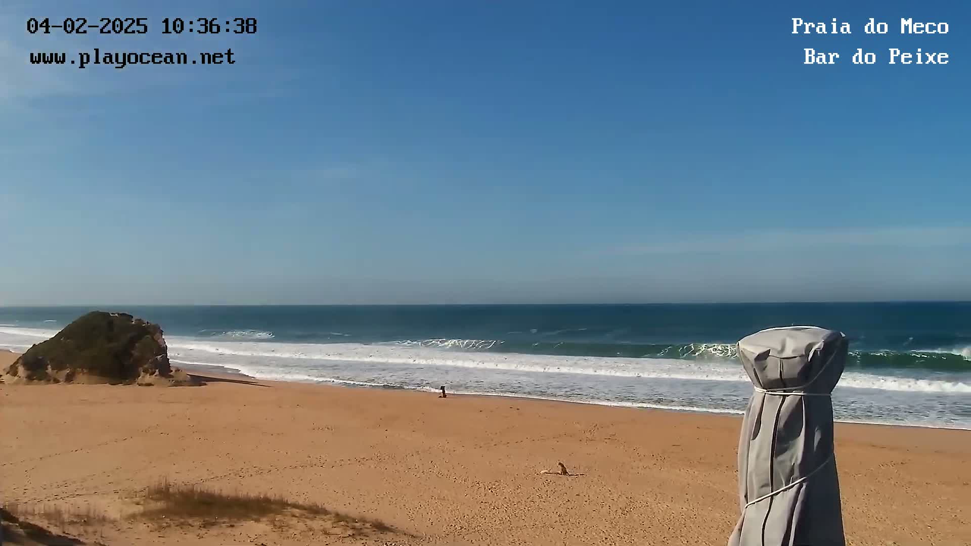 A sandy beach with small waves under a clear blue sky, and a partially covered beach umbrella in the foreground.