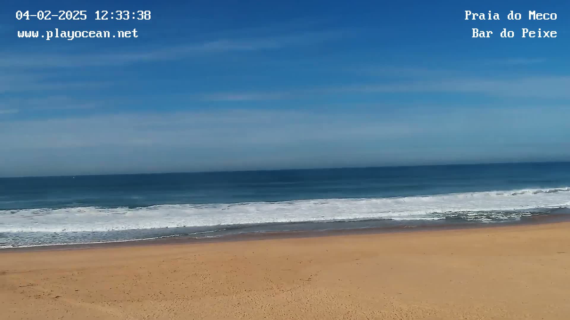 A sandy beach meets a blue ocean under a clear, mostly sunny sky with a few scattered clouds.