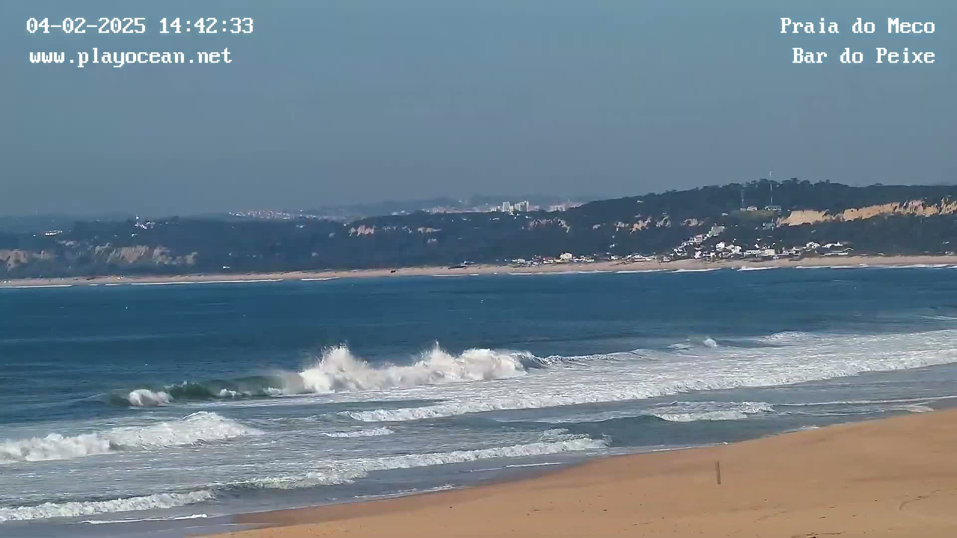 Ocean waves break on a sandy beach under a clear, bright sky, with a distant coastline visible in the background.