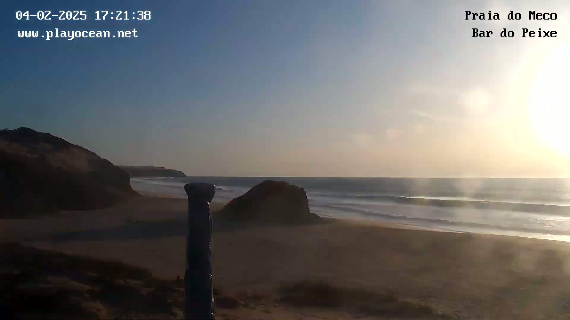 A sandy beach with small waves gently rolling onto the shore at sunset, partially obscured by a dark-colored object in the foreground.