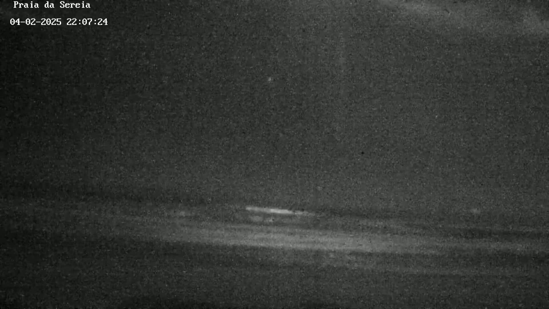 A grainy, low-light image shows a dark landscape with a faint, horizontal light source in the distance.