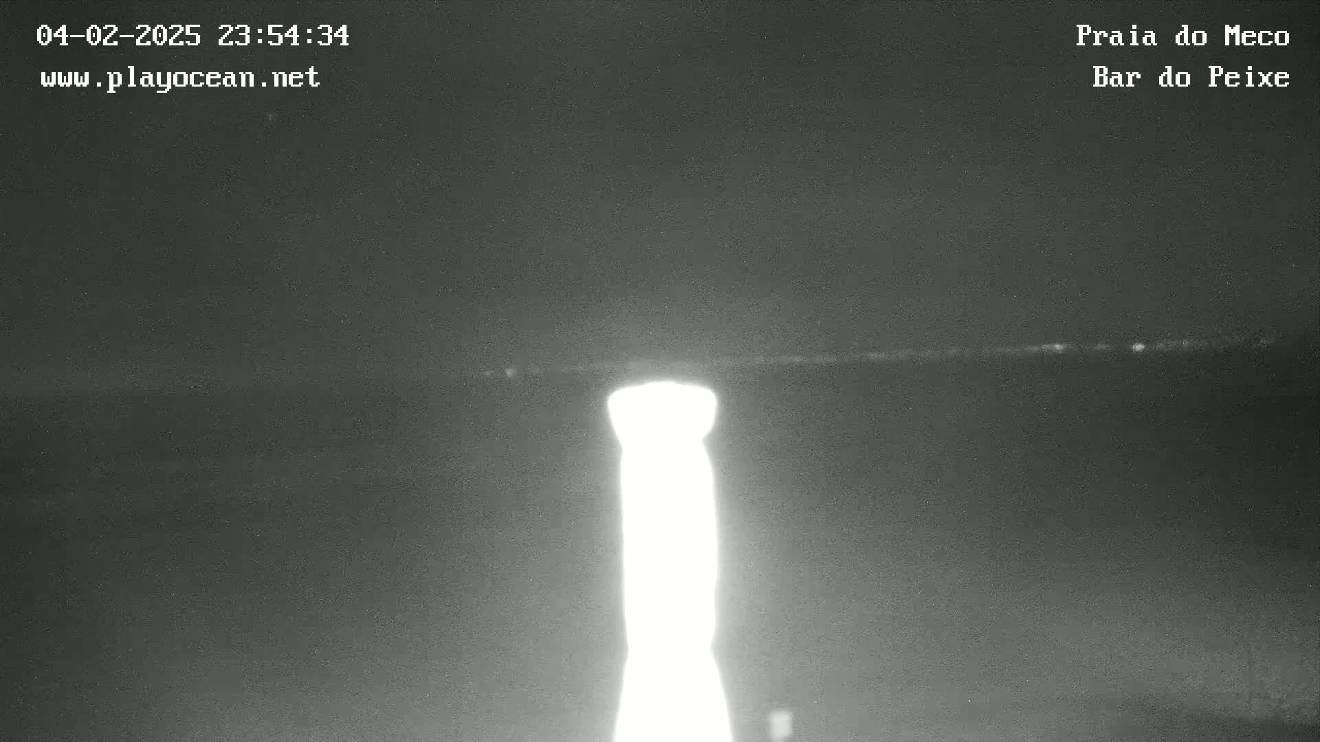 A bright light, possibly a lamp or lighthouse, illuminates the dark nighttime scene of what appears to be a beach or coastline.