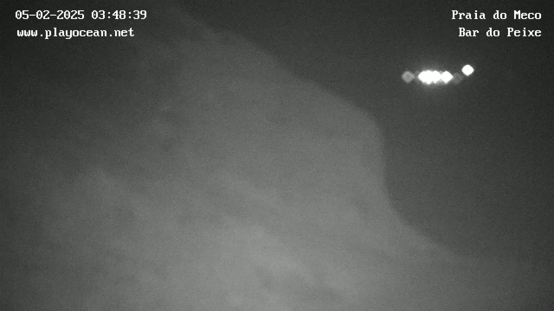 A nighttime, low-light image shows a dark, possibly sandy or beach-like area with a few distant bright lights visible.