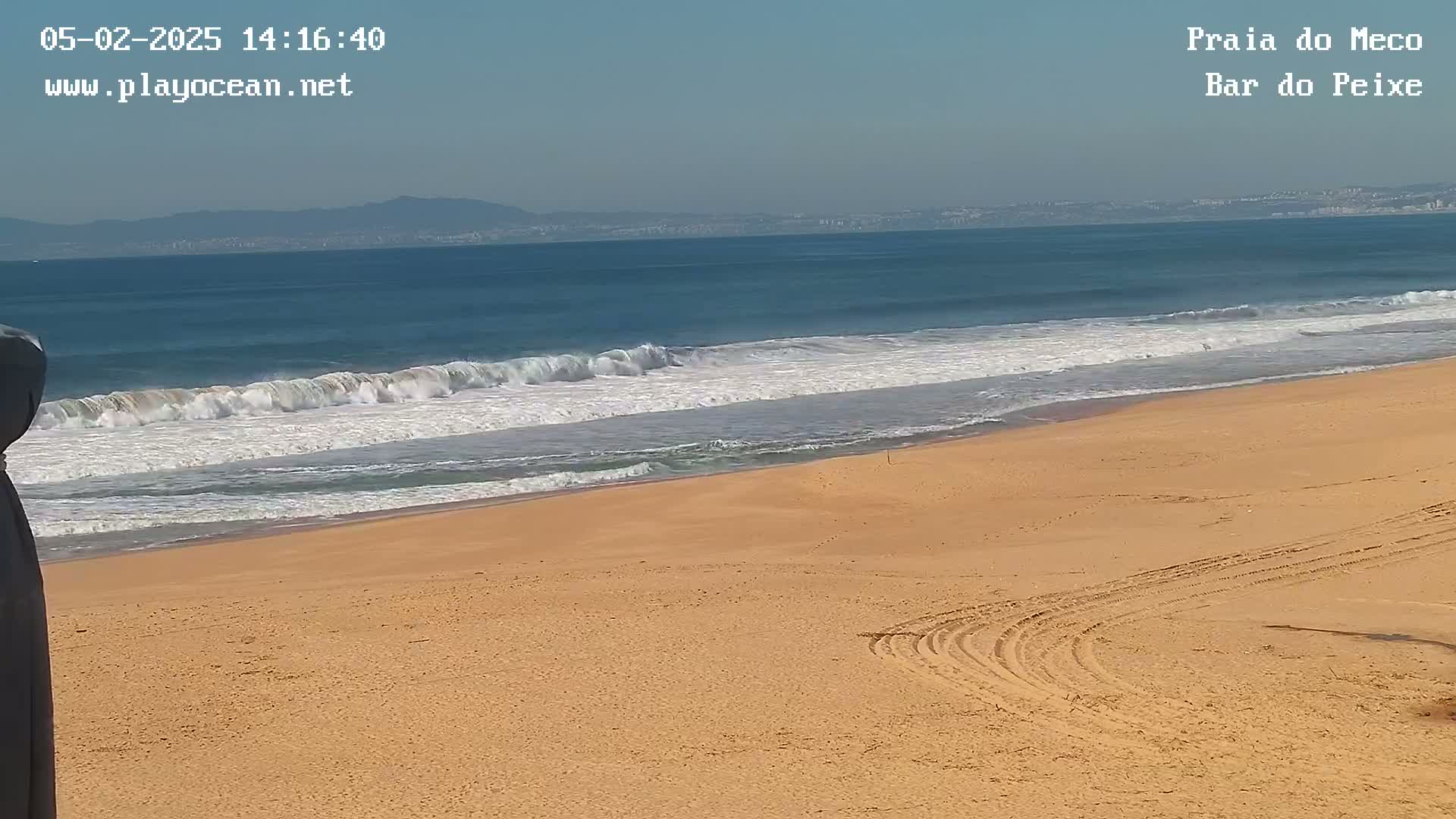 A sandy beach with tire tracks and small waves rolling onto the shore under a clear sky, with mountains visible in the distance.