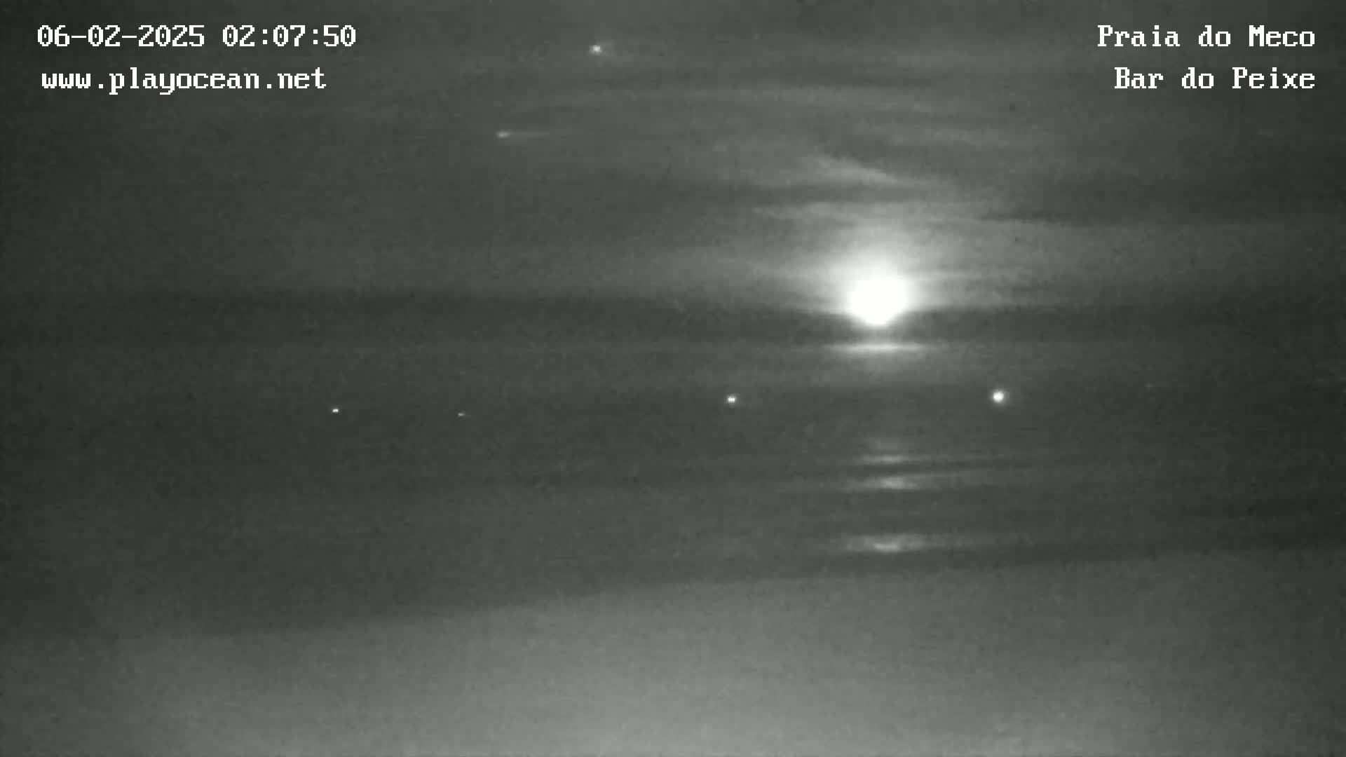 A low-light, nighttime view of a beach shows a bright moon or sun setting over calm water, with a few faint lights visible in the distance.
