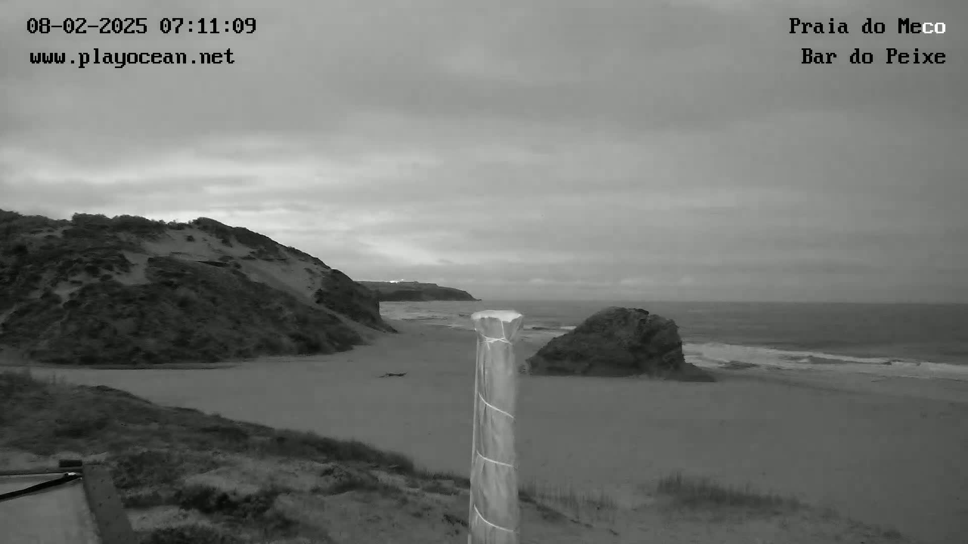 A grayscale view shows a sandy beach with a large sand dune on the left, a rock formation on the right, and a calm ocean under an overcast sky.
