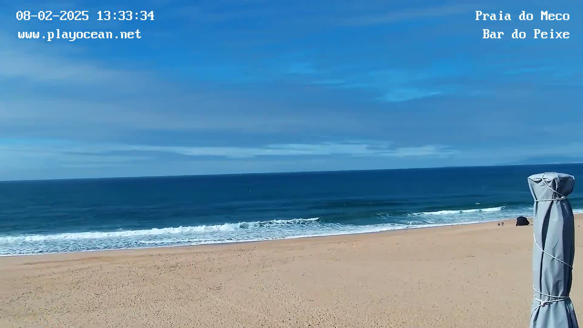 A sandy beach with gentle waves under a mostly clear blue sky is visible, with a portion of a covered beach umbrella in the bottom right corner.