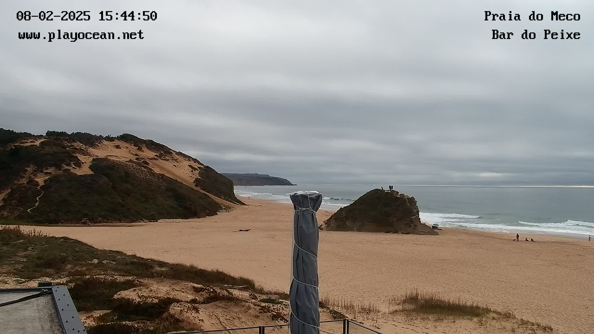 A sandy beach with low, vegetated dunes and rock formations is visible under an overcast sky, with a calm ocean in the background.