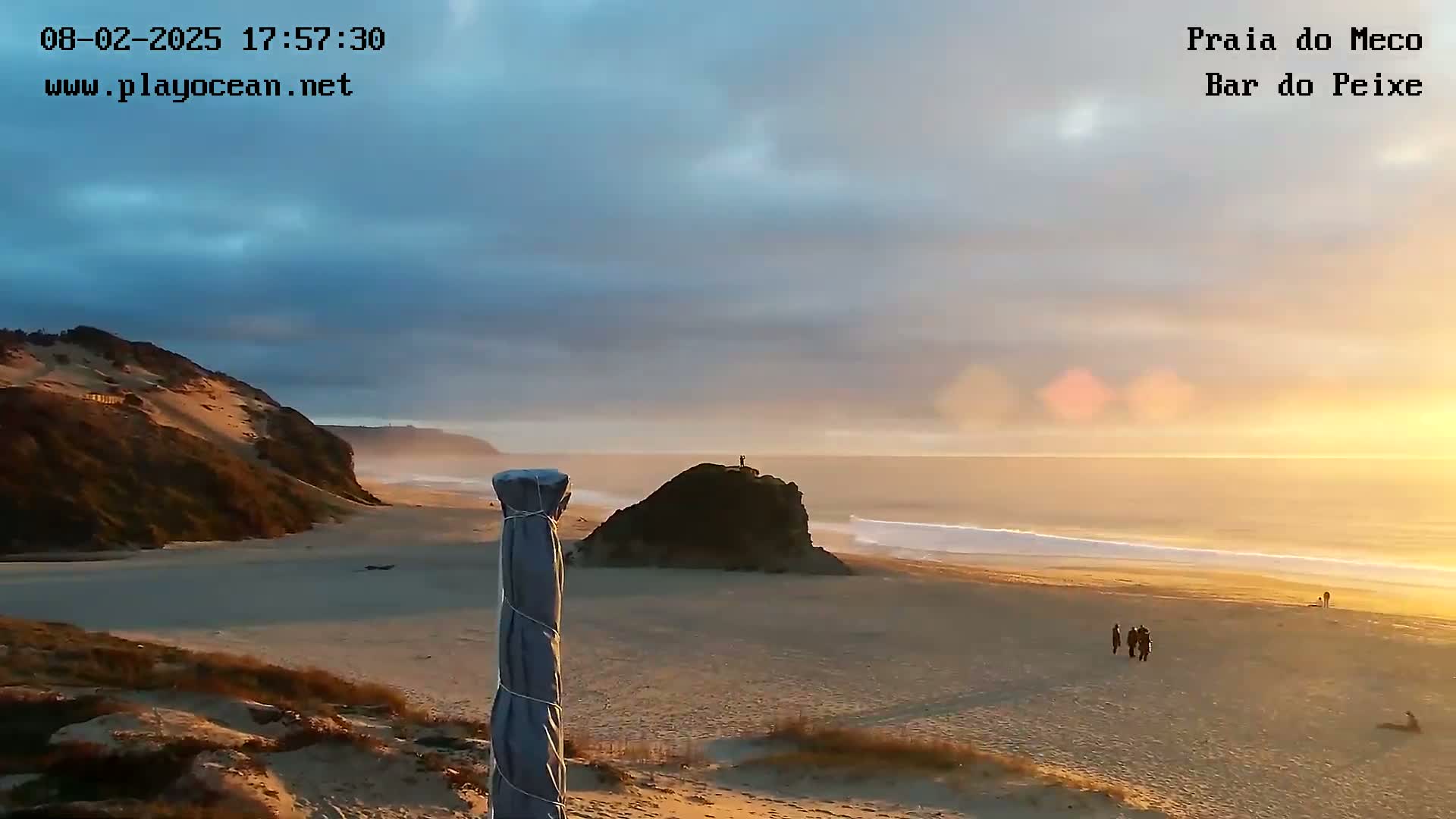 A sandy beach with a rocky outcrop and a dune-covered headland is bathed in the warm light of a sunset under a partly cloudy sky.