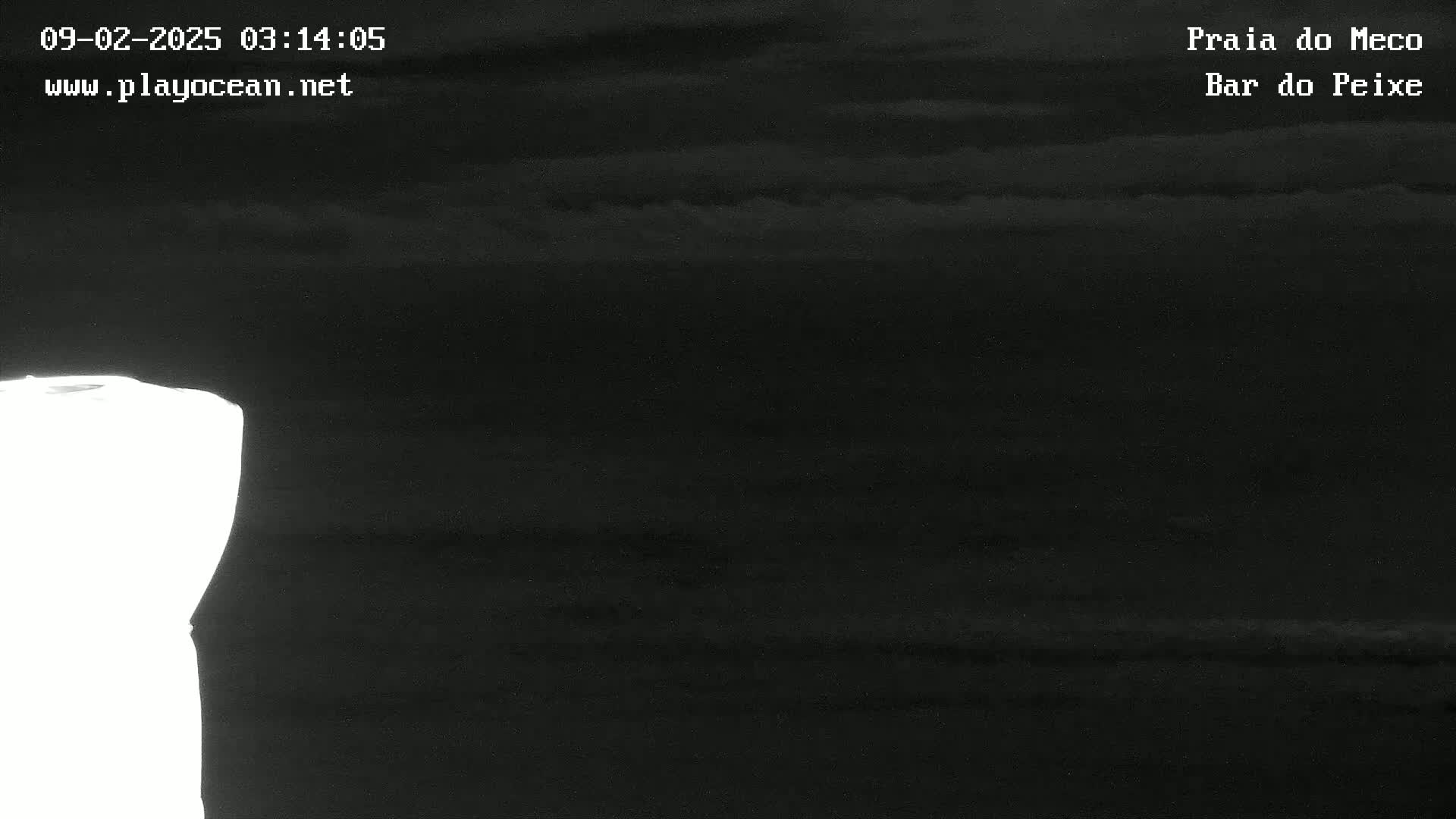 A dark, nighttime coastal scene shows a bright white area to the left, possibly a light source or reflection, against a dark, cloudy sky and dark water.